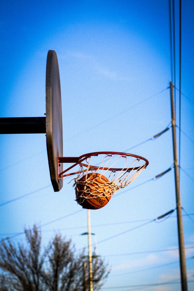 A basketball perfectly swishes through the hoop on an outdoor court against a clear blue sky.